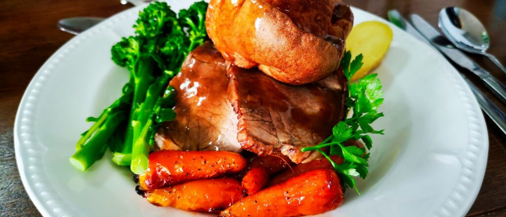 A close-up plate of roast beef with gravy, a golden Yorkshire pudding, roasted carrots, tenderstem broccoli, and a parsley garnish, set on a white plate with cutlery beside it.